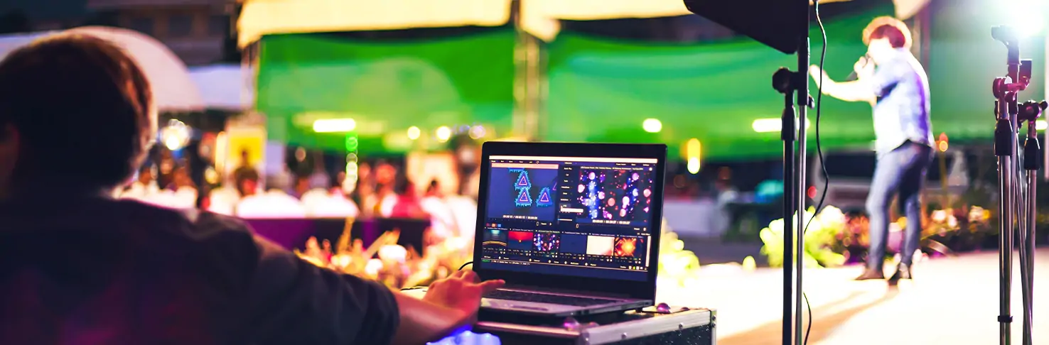 View from behind a stage technician working on a laptop with event production software, overseeing a live performance on a secure outdoor stage in Panama City, Florida. The vibrant atmosphere is captured with colorful stage lighting and an engaged audience in the background, illustrating Neves Media's comprehensive stage services that ensure a seamless event experience.