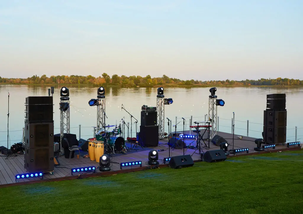 An outdoor stage set by the serene waters in Panama City, equipped with professional audio and lighting gear. The tranquil setting is enhanced by the safe and sturdy stage platform, showcasing a ready-to-perform music setup with various instruments, reflecting the high-quality and reliable rental services available for any waterfront event.